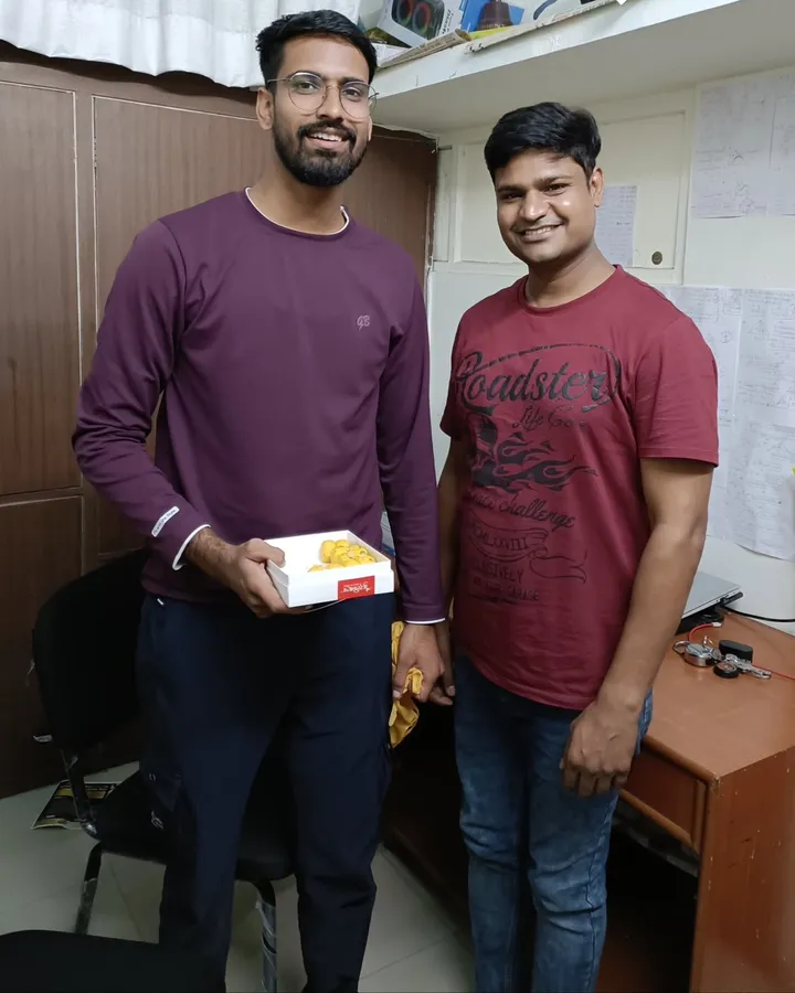 Akshay Khapre and a member of the Vivekananda team standing together in the institute office, a box of sweets between them, after his Sub Inspector selection
