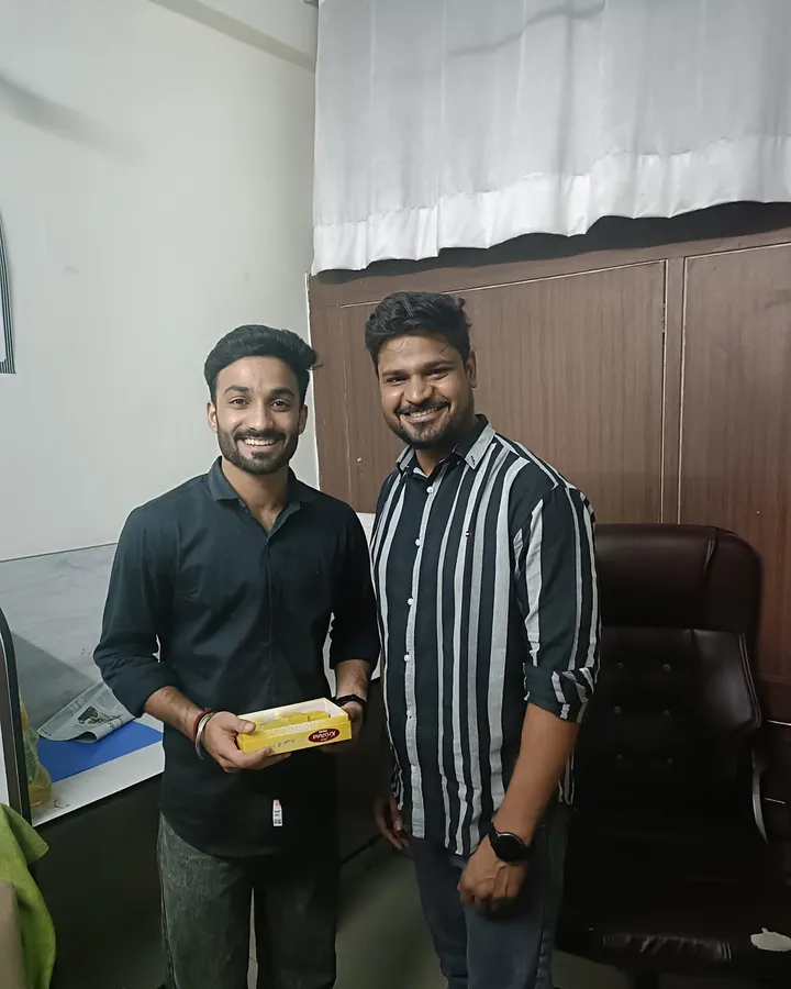 Himanshu with a box of sweets, standing with a member of the Vivekananda team in front of the office cupboards after his CAT result