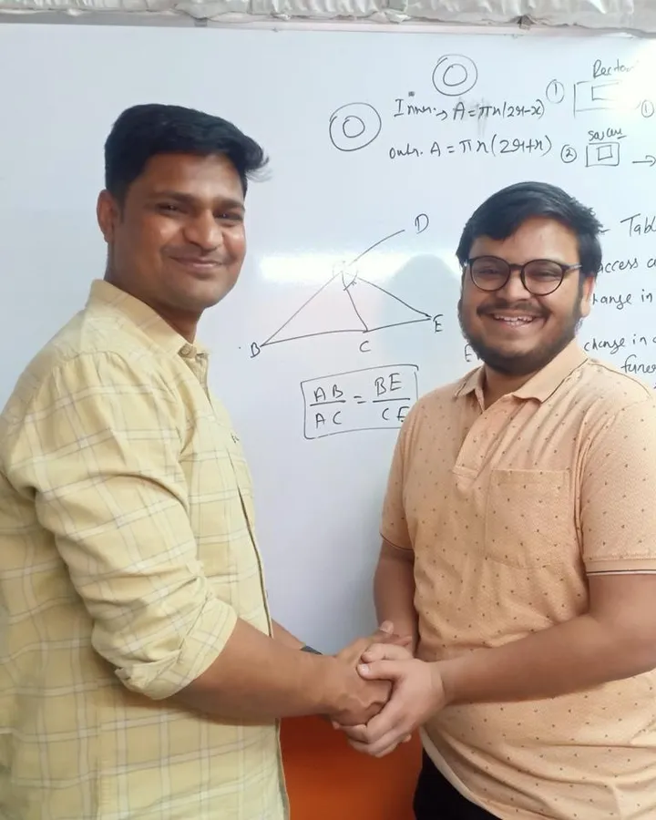 Saksham Bajaj shaking hands with a member of the Vivekananda team in front of a whiteboard covered in worked problems, after his GATE result