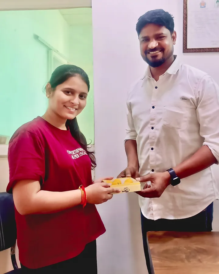 Sneha Patil being handed a plate of sweets at the institute, framed certificates on the wall behind her, after her IBPS selection