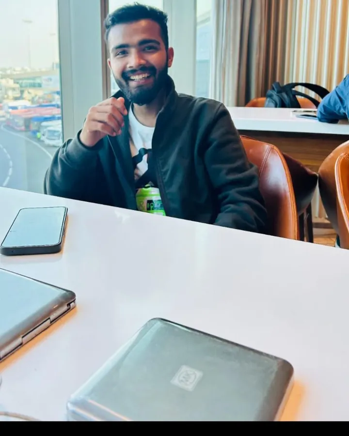 Samyak Jain giving a thumbs up at a table by a window, a laptop and phone in front of him, after qualifying as a CA