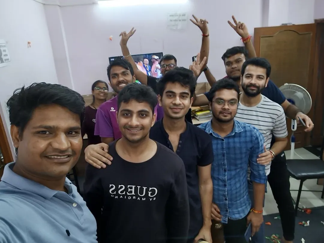 A group of students packed into frame for a selfie, arms raised, at a gathering indoors