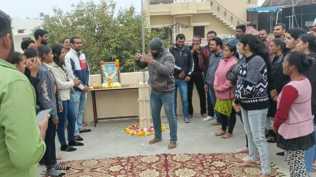 Students standing in a circle on carpets on the terrace around a garlanded portrait of Saraswati and a marigold rangoli, watching a lamp being lit