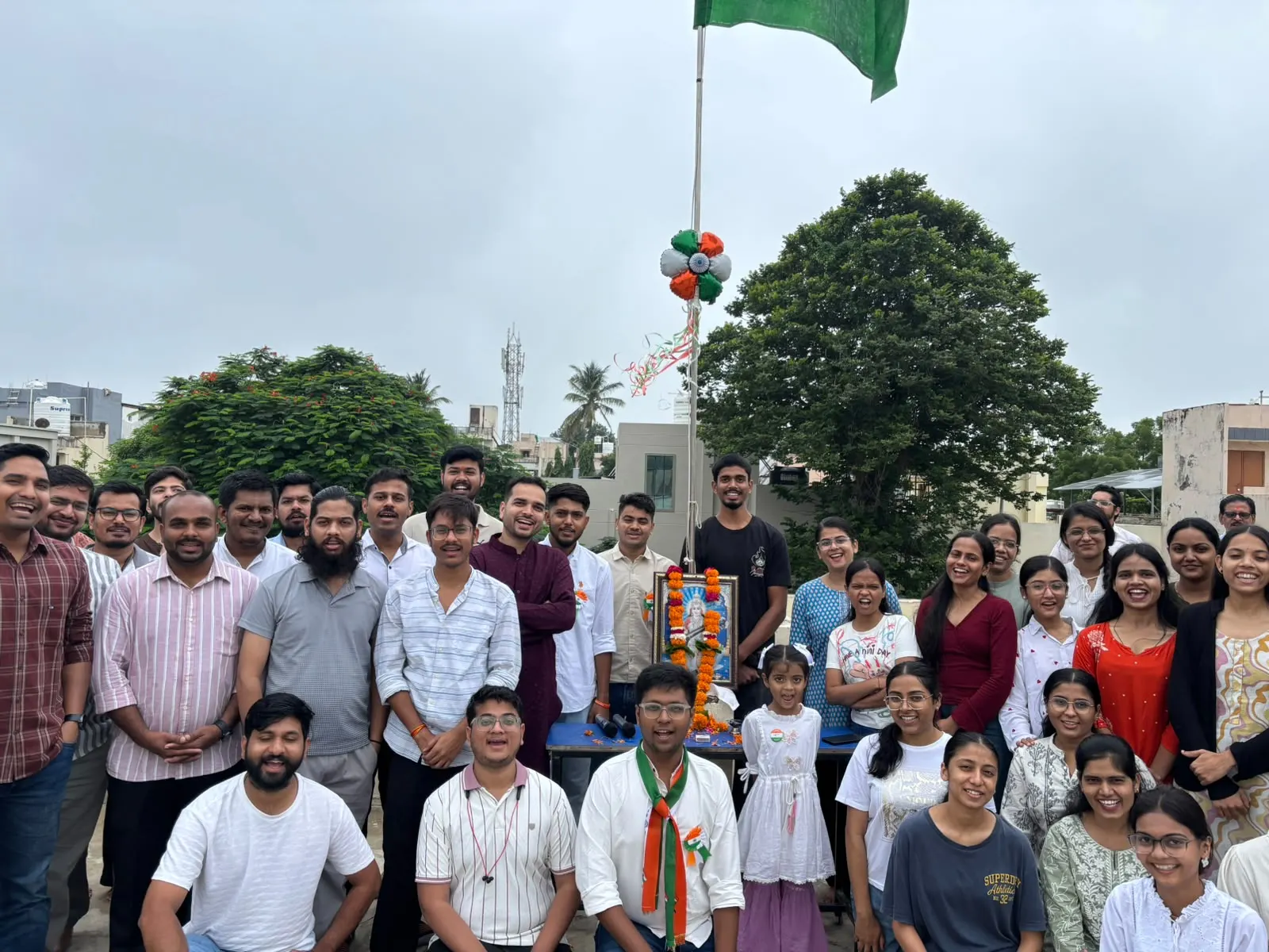 Students and staff gathered on the terrace around a flagpole hung with tricolour balloons and a garlanded portrait