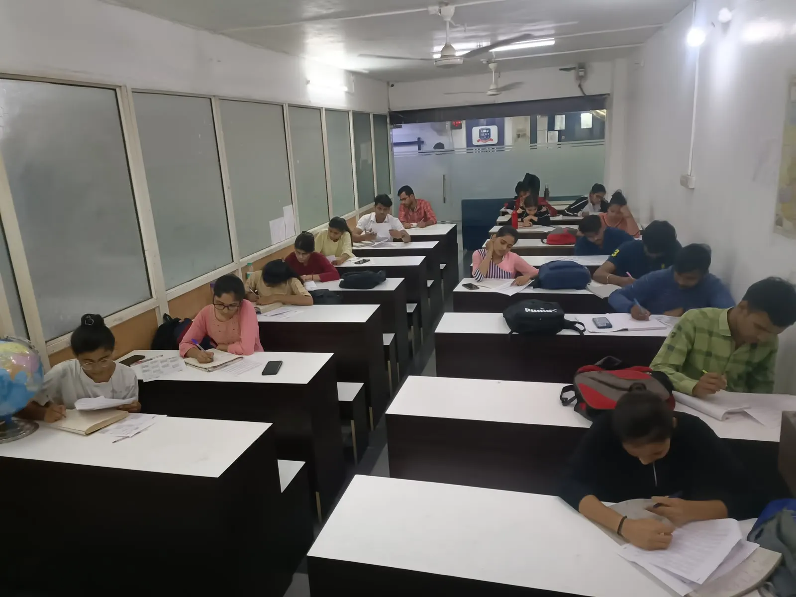The study hall in use: rows of individual desks with high partitions, each student working alone under strip lighting, with bags and water bottles beside them
