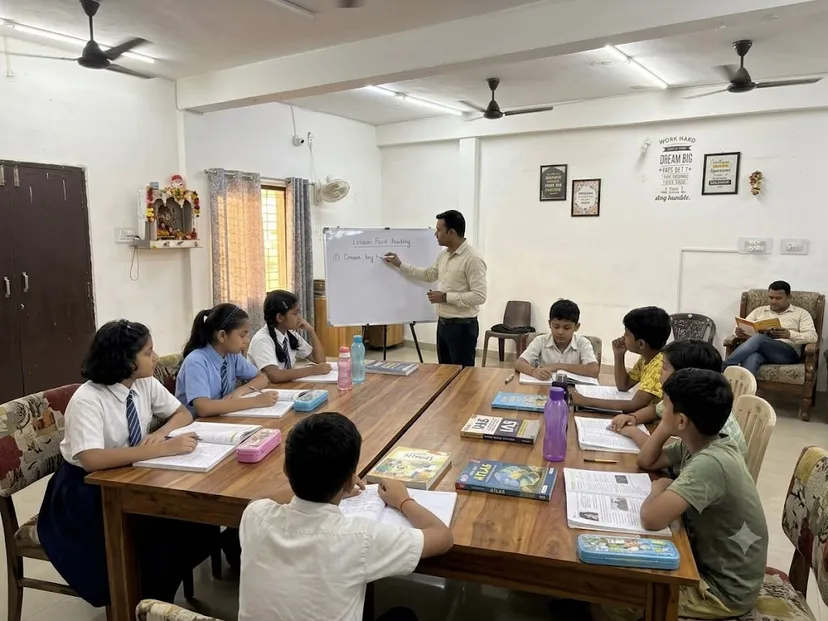 A teacher at a whiteboard with school children in uniform seated around a shared table, textbooks and pencil cases open in front of them